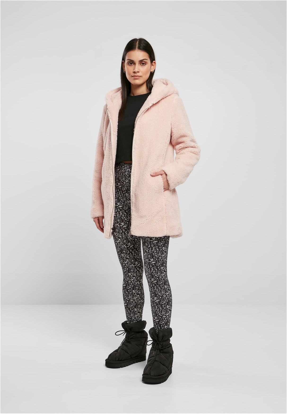 En kvinna visar framsidan av en rosa sherpajacka med huva, buren med mönstrade leggings och svarta stövlar.