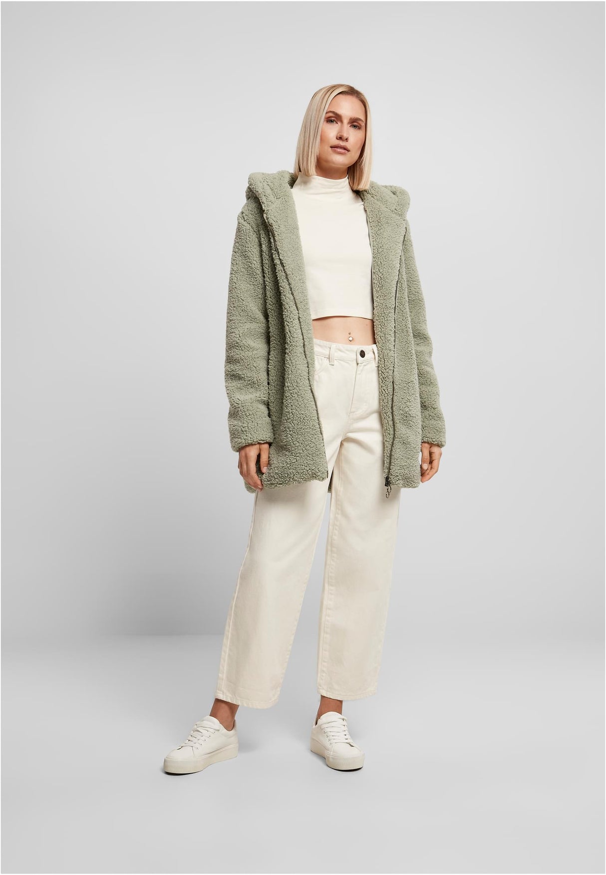 En kvinna bär en grön, öppen UC Sherpa Jacket med huva, tillsammans med en vit topp och vita byxor.