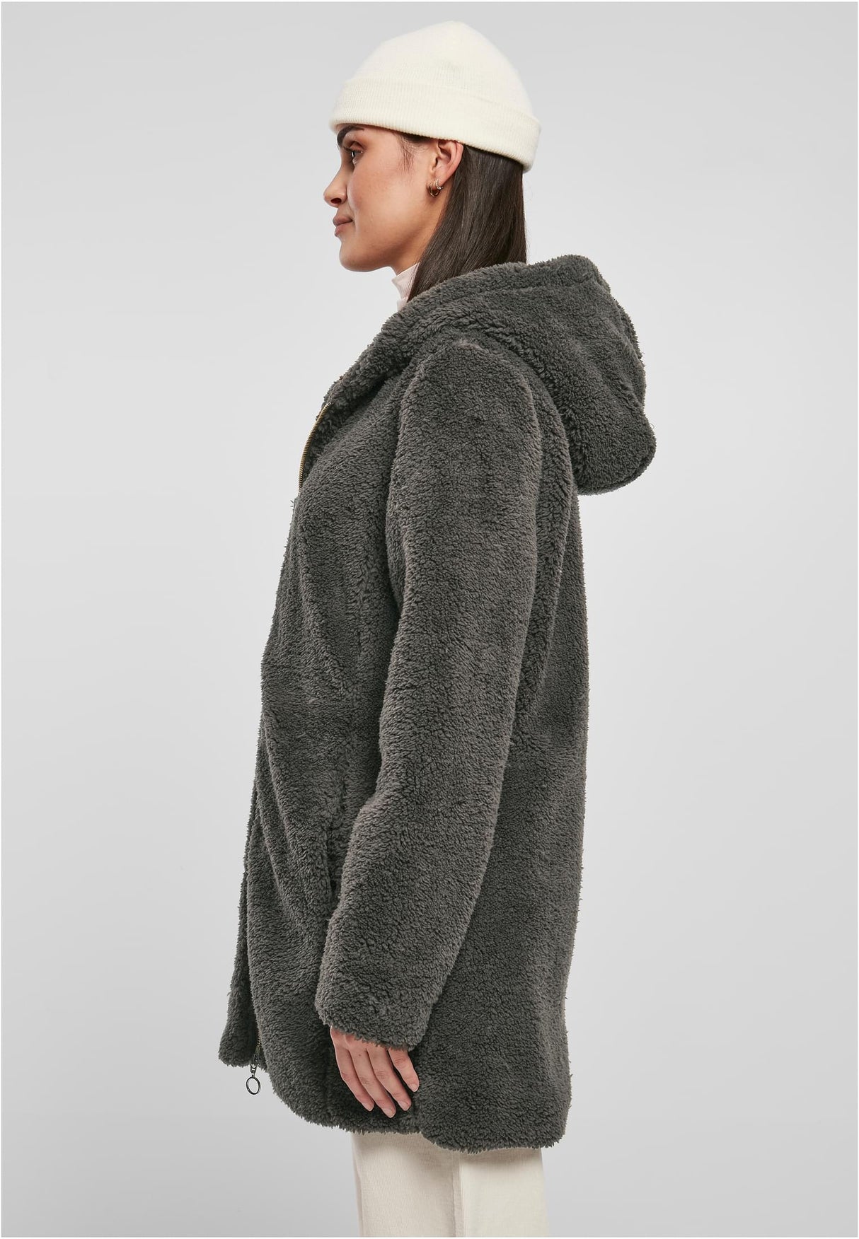 En kvinna sedd från sidan som bär en mörkgrå, luddig sherpajacka med huva och en vit mössa.