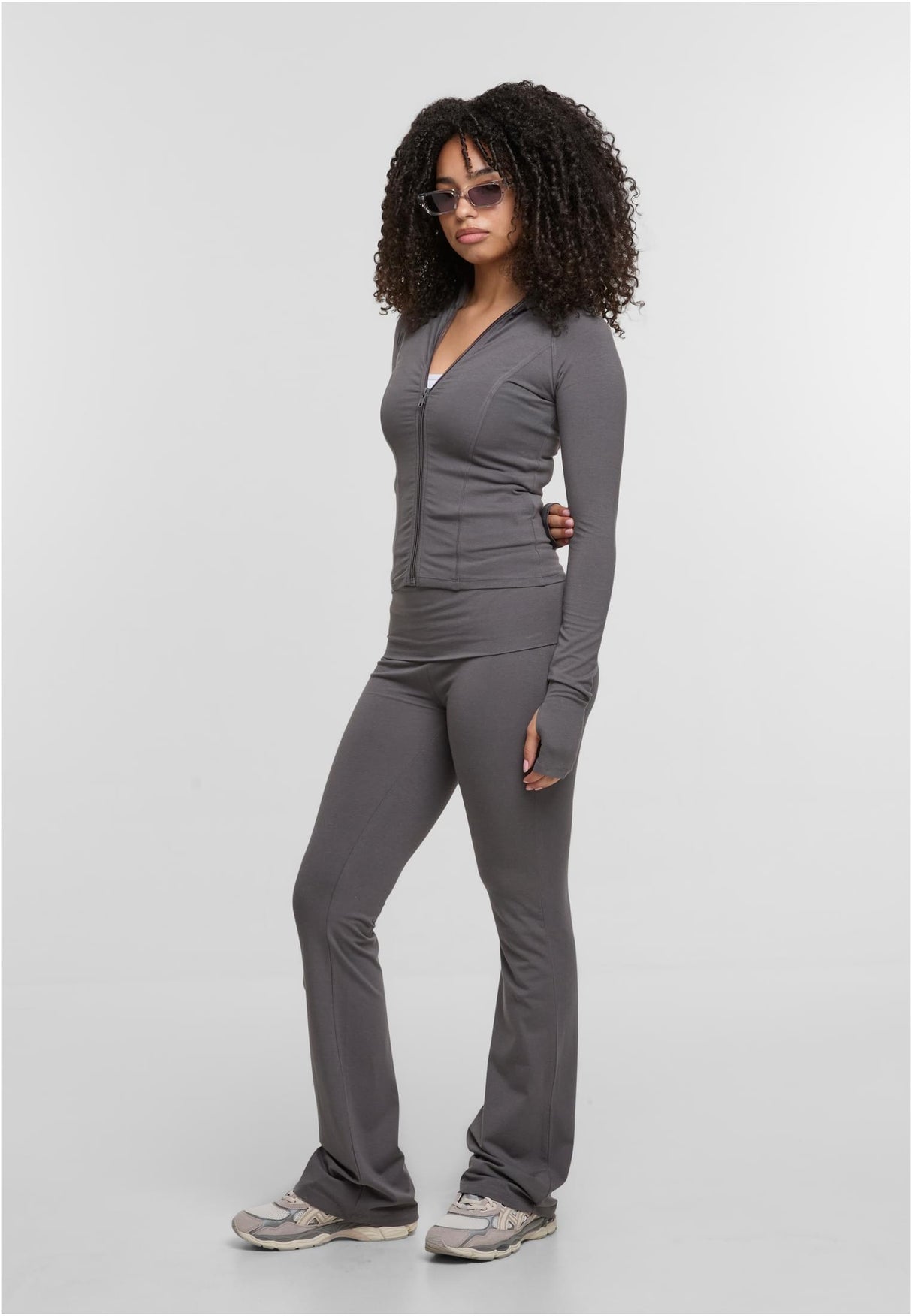 En kvinna poserar i en grå träningsjacka med dragkedja och matchande utsvängda byxor mot en vit bakgrund.