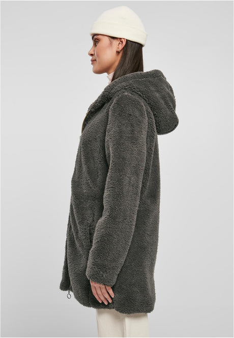 En kvinna sedd från sidan som bär en mörkgrå, luddig sherpajacka med huva och en vit mössa.